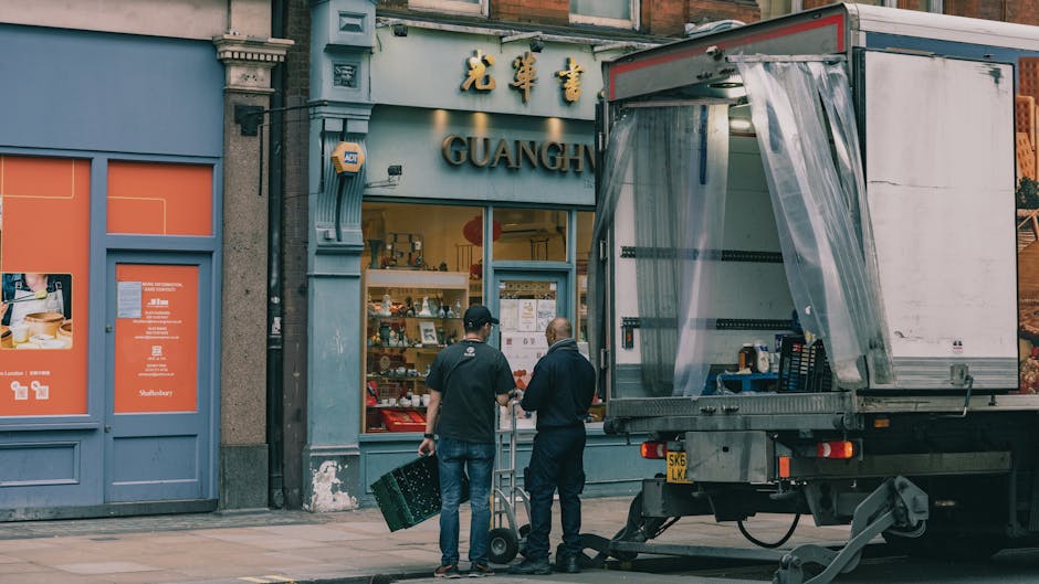 Two removal workers from Belgravia Removals are preparing to load moving boxes onto a delivery truck outside a commercial premises in Belgrave Square, London. The boxes are made of cardboard and are positioned on the pavement near the truck's open side, which is partially covered with a transparent plastic sheet. One worker, wearing dark clothing, stands next to a hand truck, while the other, dressed in a black shirt and jeans, carries a small crate. The background features the storefront of a shop with signage in Chinese characters and the name 'GUANG' in English, and the shop window displays various products. The scene is set during daylight hours, with natural light illuminating the area, and the surroundings suggest a typical busy city street engaged in home relocation or furniture transport activities, as part of the packing and moving process supported by Belgravia Removals.