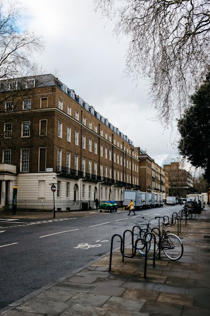 A street scene outside a multi-storey brick building with white window frames and black balcony railings, capturing a home relocation process involving furniture and boxes. A large blue moving truck is parked along the curb directly in front of the building, with its tailgate lowered for loading or unloading. Several furniture pieces covered with blankets and secured with straps are being moved, visible near the entrance, where a worker is handling equipment such as trolleys and moving straps. The sidewalk has a row of empty bike racks, with a few bicycles parked nearby, while the street is lined with parked cars and bicycles. Overhead, leafless tree branches extend across the scene, and the sky appears cloudy, indicating typical urban conditions. The presence of residential windows, street signage, and pavement markings situates this within a city environment suitable for detailed descriptions of house removals and furniture transport logistics, as offered by Belgravia Removals.