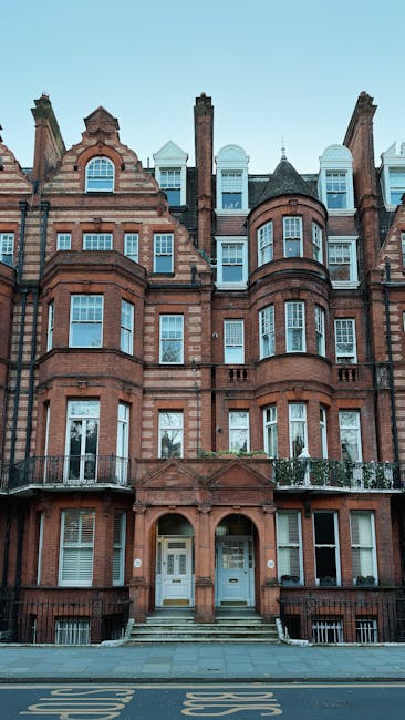 A multi-storey red brick Victorian-style residential building with ornate architectural details, including arched windows and decorative stonework, situated on a narrow street with a sidewalk and a bus stop in front. The building features several balconies with metal railings, some with potted plants, and a central entrance with steps leading up to white double doors. The clear sky above and the street below indicate a daytime setting. This image visually relates to house removals and furniture transport, illustrating the type of property typically serviced by Belgravia Removals during home relocation or moving processes.