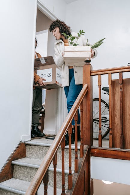 A cheerful man, dressed in a maroon t-shirt and orange trousers, is standing on a staircase while carrying a medium-sized cardboard box with red tape, labeled with handling instructions and contents, 
